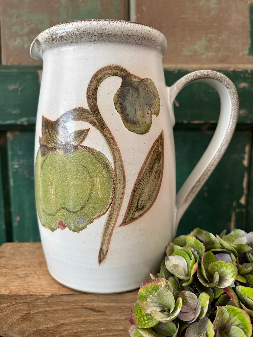 A white Denby ceramic pitcher with a rustic speckled rim and handle, decorated with a green and brown botanical illustration of a flower and leaves. 