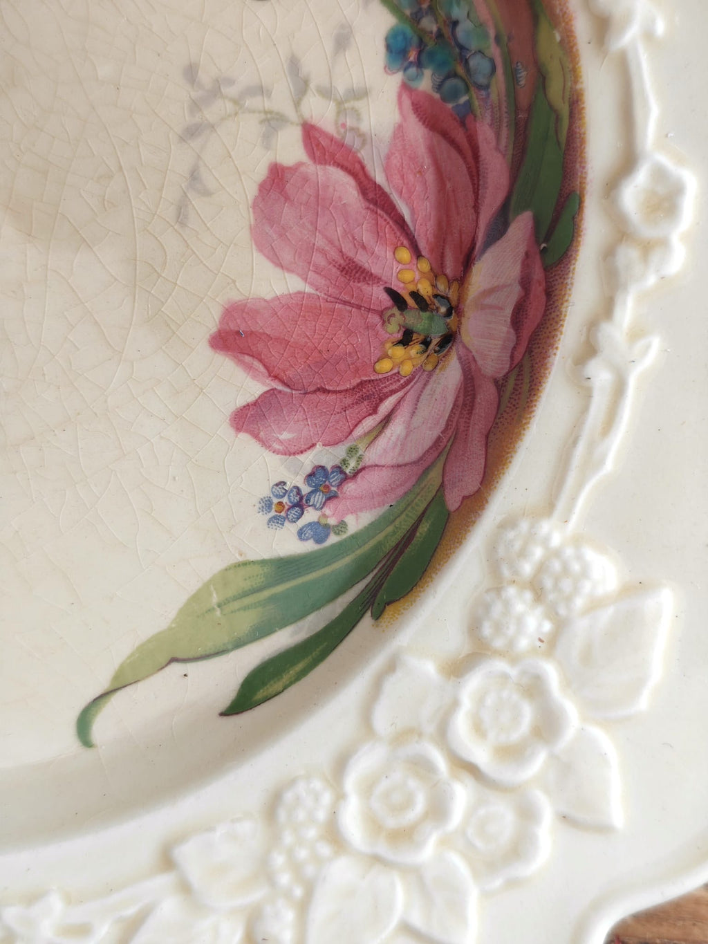 Close-up of a vintage white ceramic plate with a pink flower and green leaves painted on the inside, featuring embossed floral details along the rim.