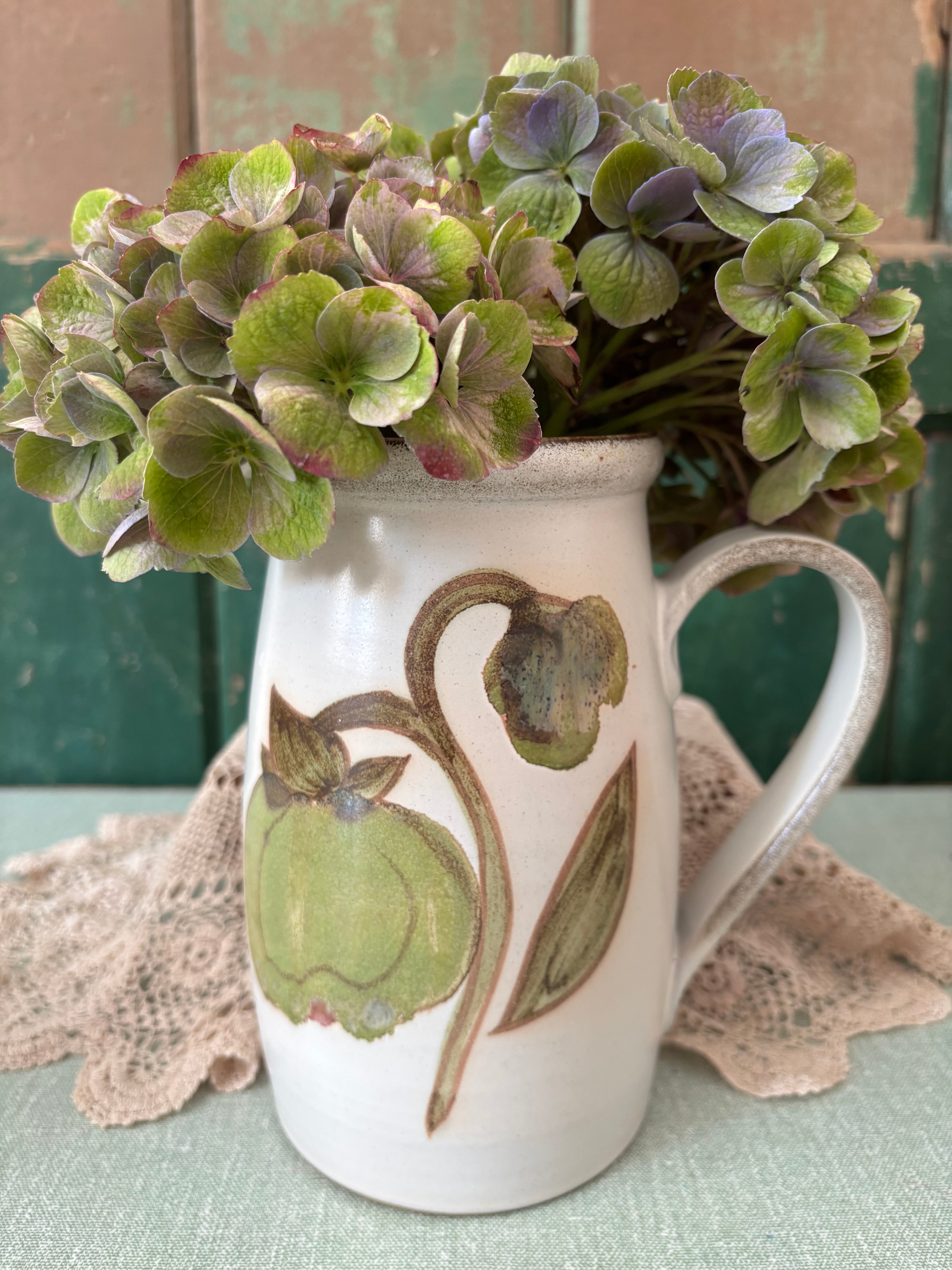 A white ceramic pitcher with a rustic handle and a painted green botanical design.