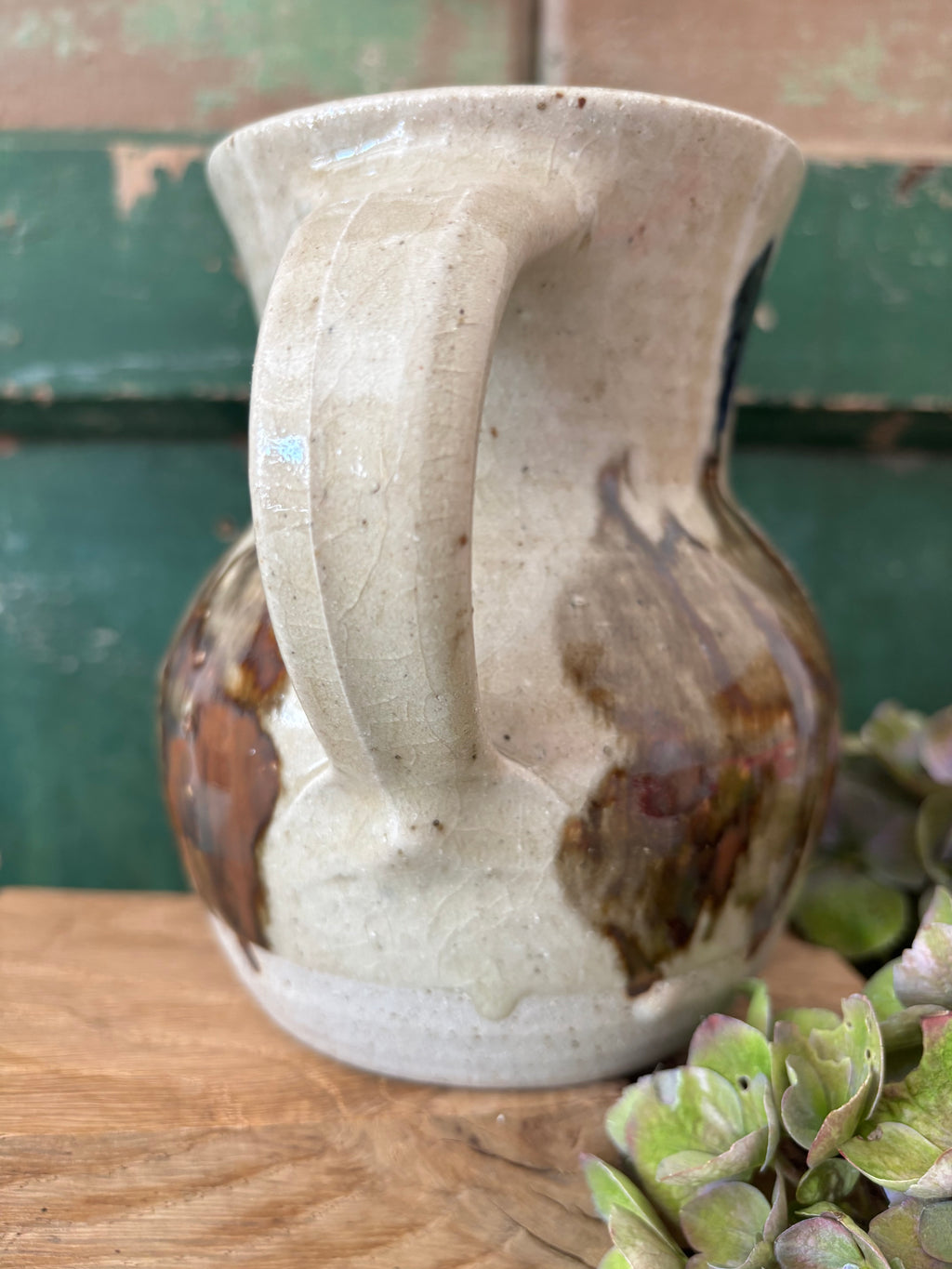 A handmade ceramic pitcher with a rounded body, ribbed handle, and wide flared rim, featuring a speckled off-white glaze with brown and dark amber marbling. 