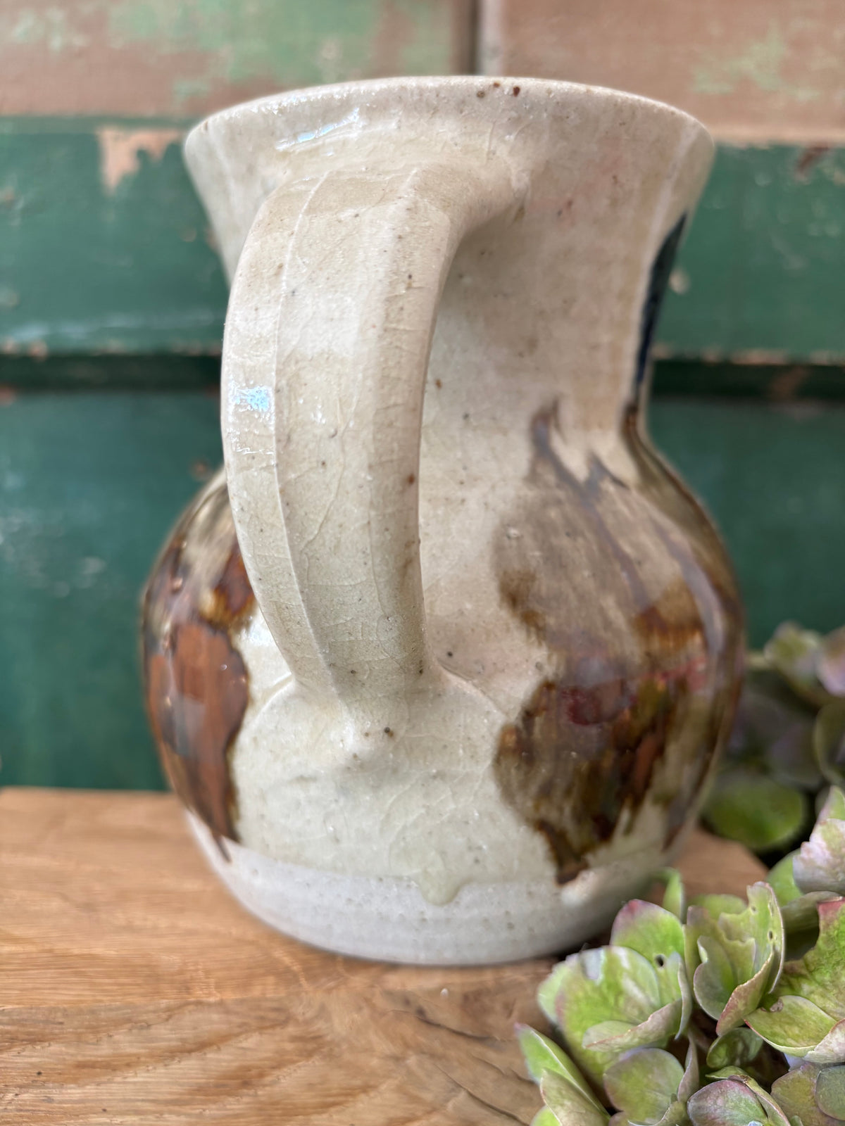 A handmade ceramic pitcher with a rounded body, ribbed handle, and wide flared rim, featuring a speckled off-white glaze with brown and dark amber marbling. 