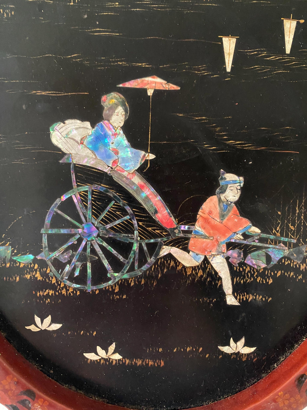 Detail of a decorative panel with a scene of a woman in a rickshaw being pulled by a man, on a black background.