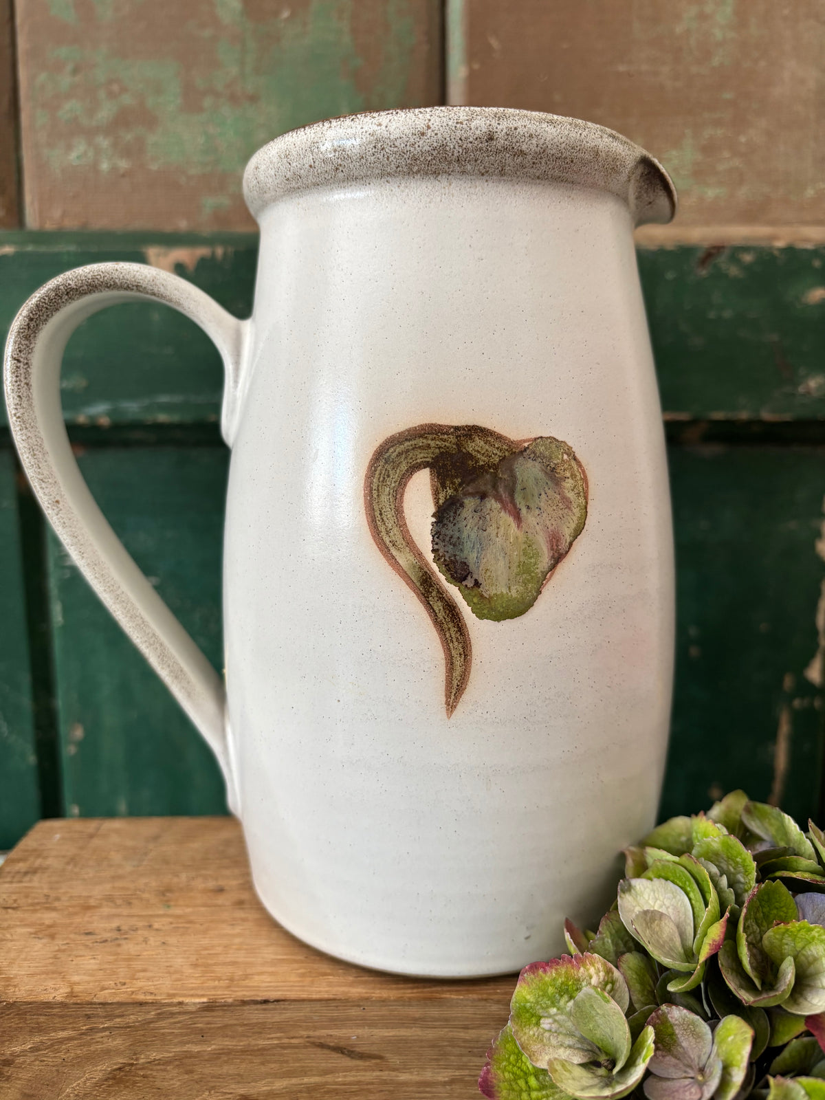 A white ceramic pitcher with a rustic speckled rim and a small painted bud design in earthy green and brown tones.
