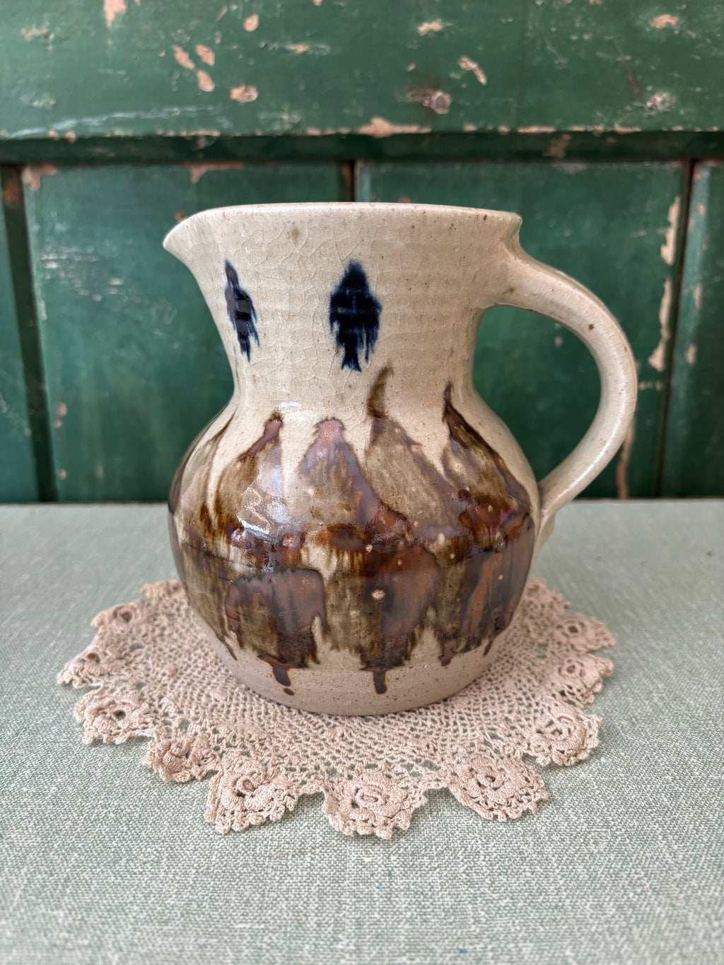 A handmade ceramic pitcher with a cream-colored glaze, dark blue leaf-like patterns near the neck, and brown dripped glaze accents on the body.