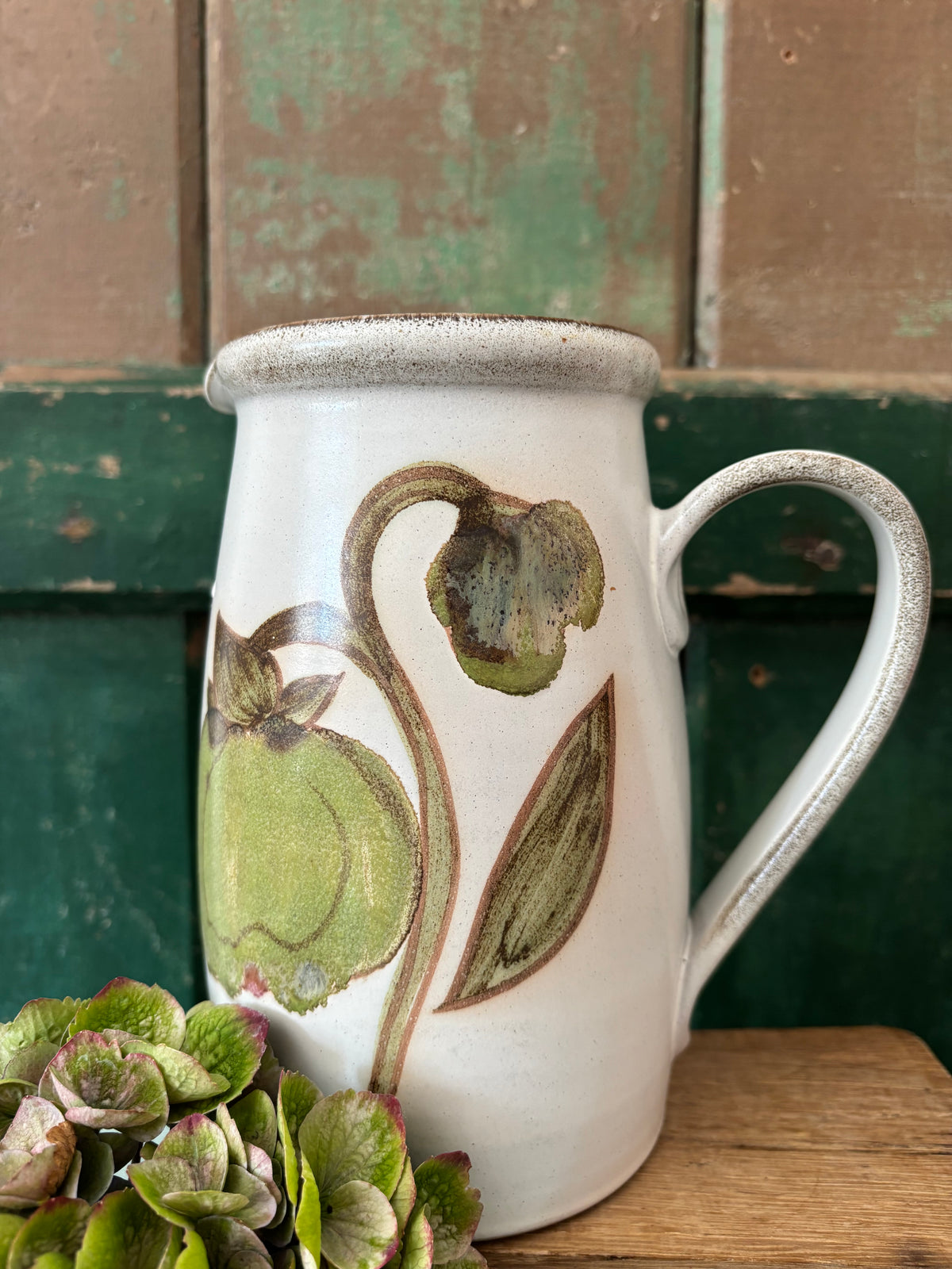 A white ceramic pitcher with a rustic, speckled rim and handle, decorated with a painted green magnolia bud motif. 