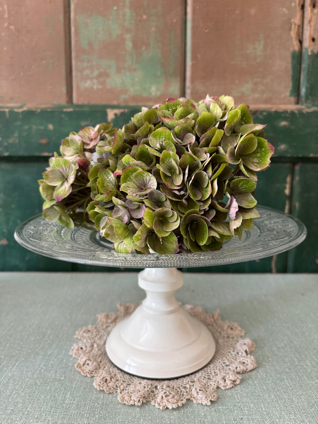  A clear glass pedestal cake stand with a cream base and a lace doily.