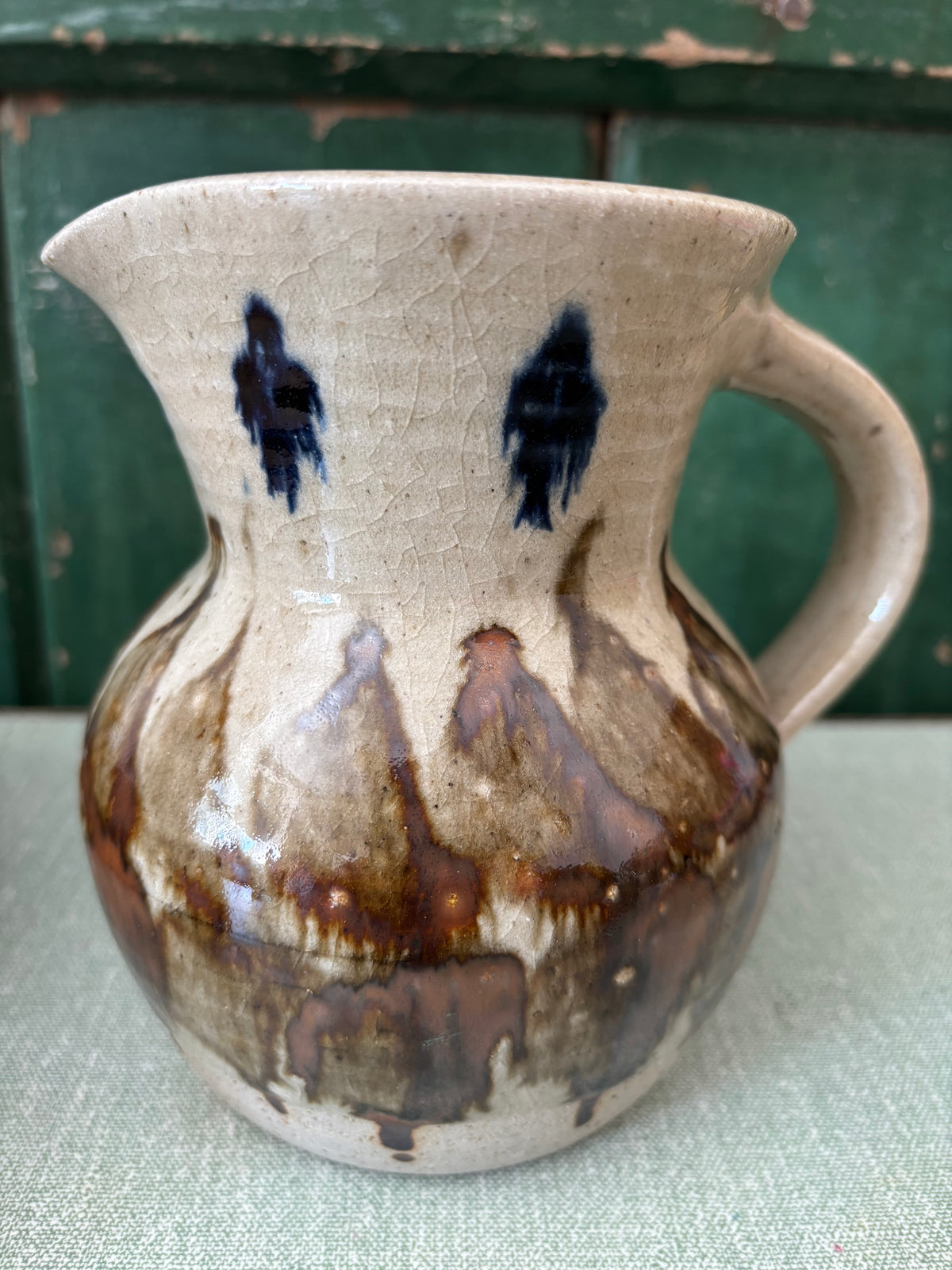 A handmade ceramic pitcher with a cream-colored glaze, dark blue abstract speckles near the rim, and a brown and beige drip pattern on the body.