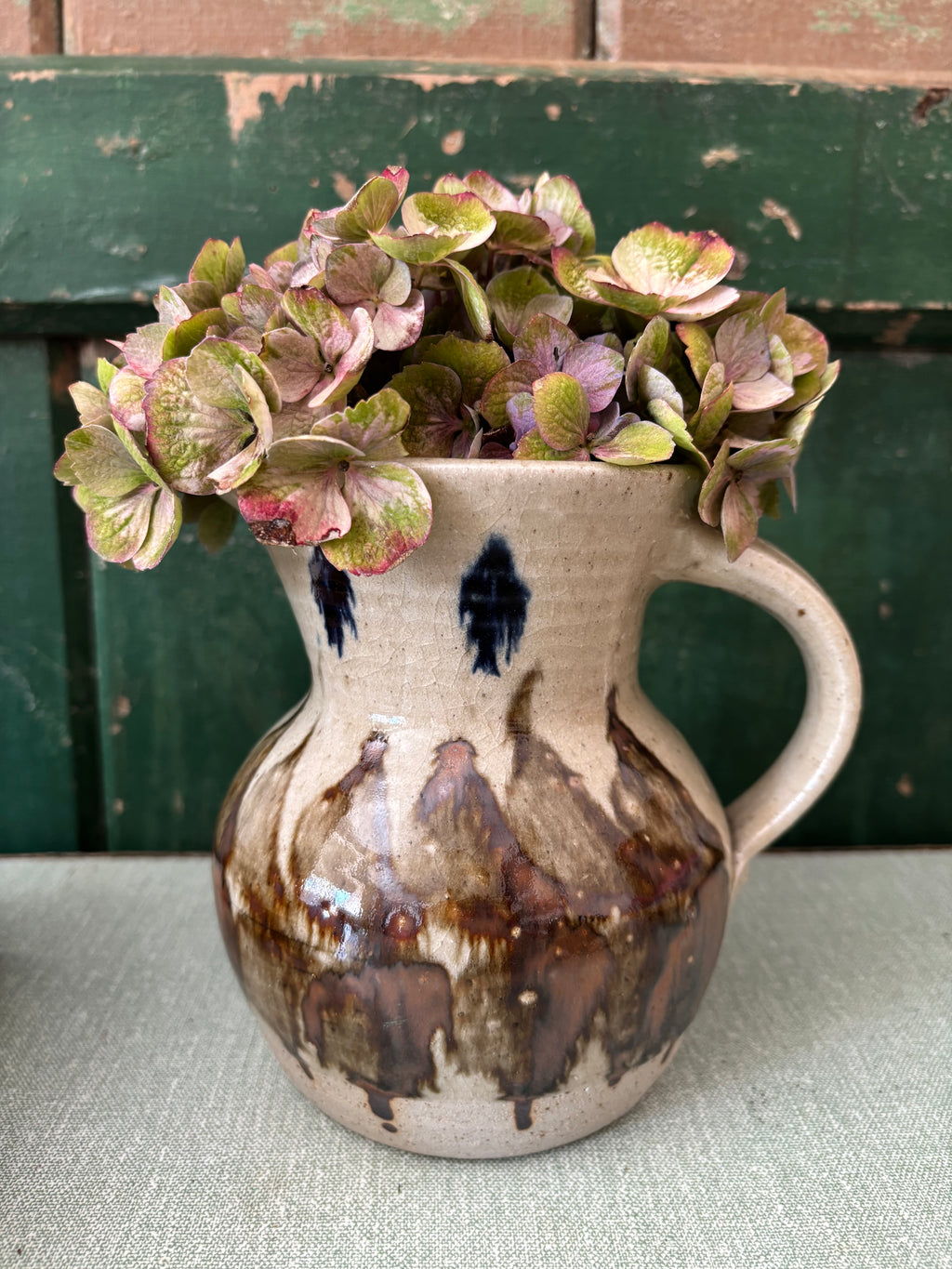 A handmade ceramic pitcher vase with a beige base, dark blue speckles, and brown glaze drips.