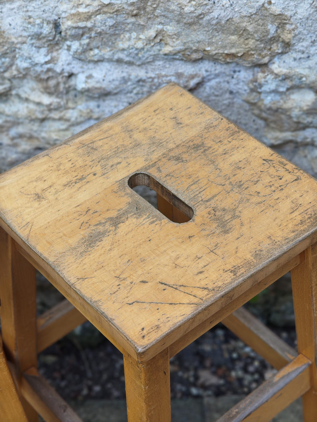 A rustic wooden stool with a square seat featuring a central cutout handle