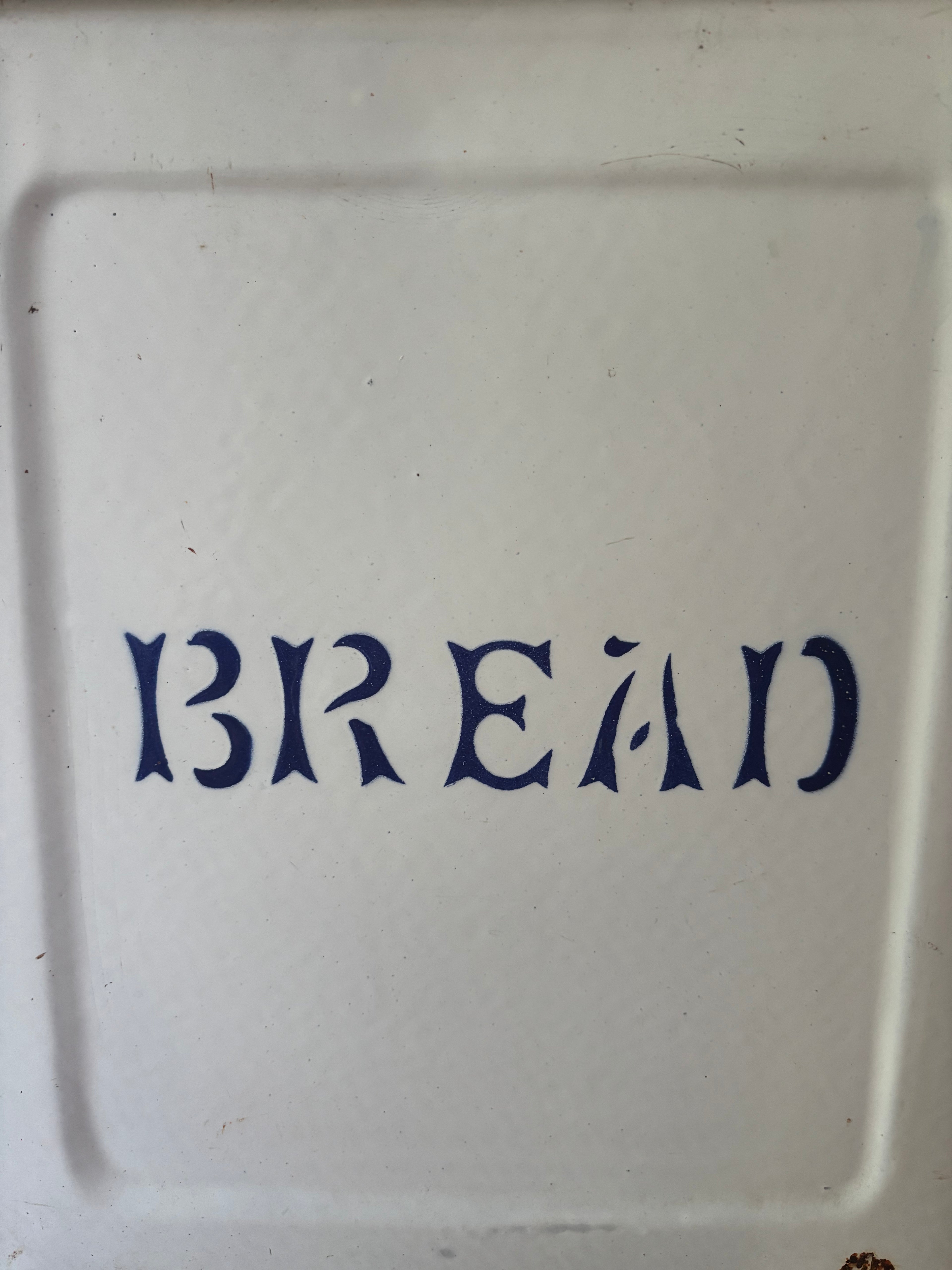 A close-up of a white enamel bread tin with the word 'BREAD' printed in dark blue.