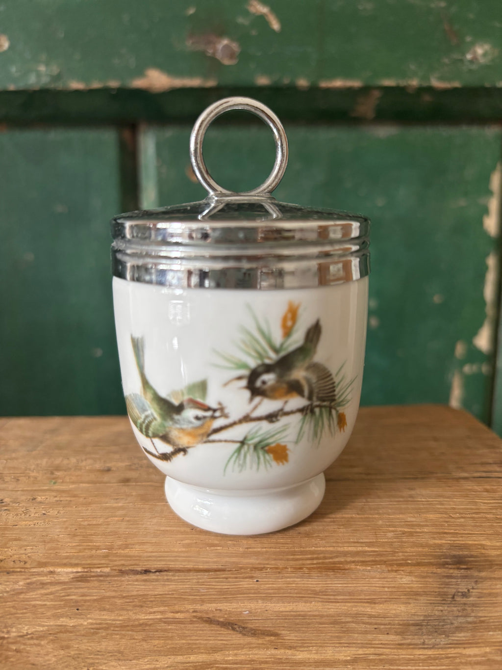 A vintage white ceramic lidded container with a silver metal ring handle and silver band, decorated with a painted bird and pine branch motif.