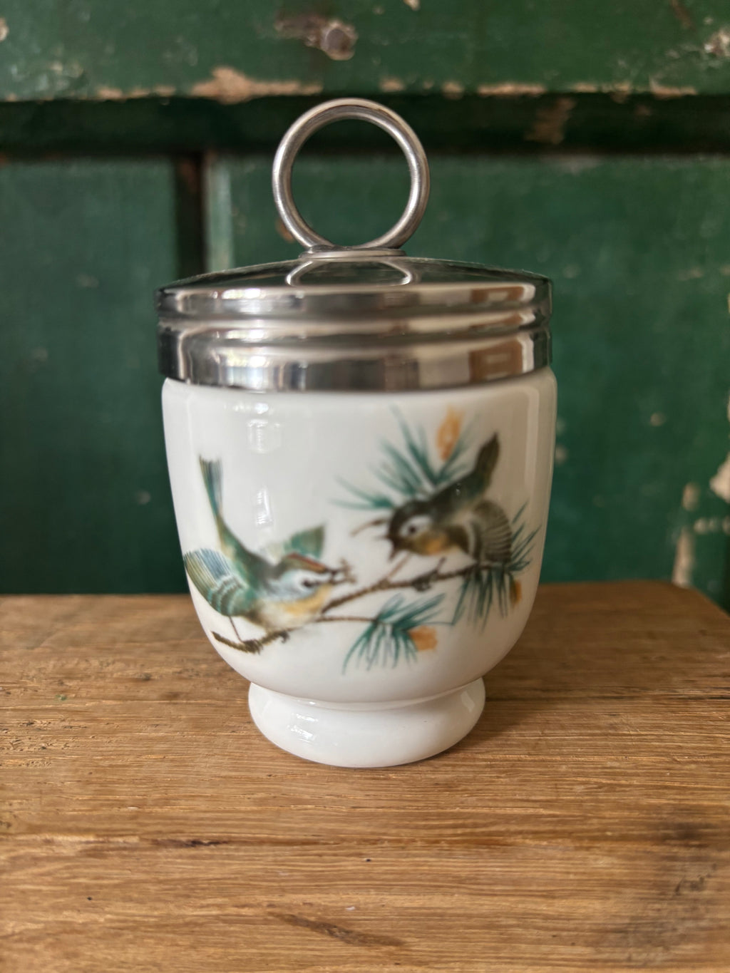 A vintage white ceramic lidded egg coddler with a silver metal ring handle and lid, decorated with a painted bird and pine branch motif.