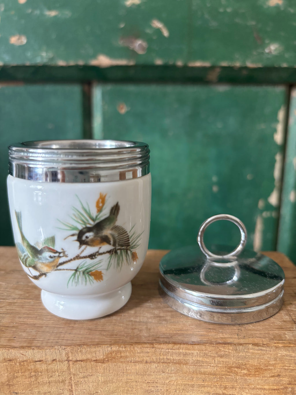 A vintage white ceramic egg coddler with a silver metal lid, decorated with a painted bird design.