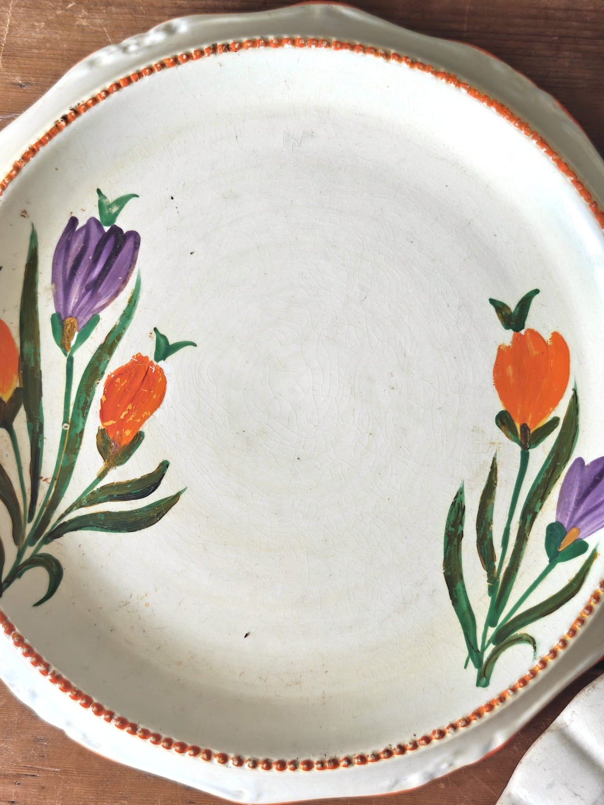 A round off-white ceramic serving platter with scalloped edges, decorated with two painted floral clusters in purple and orange with green stems, and a thin orange beaded border. 