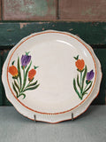 A round off-white ceramic serving platter with scalloped edges, decorated with two painted floral clusters in purple and orange with green stems, and a thin orange beaded border. 