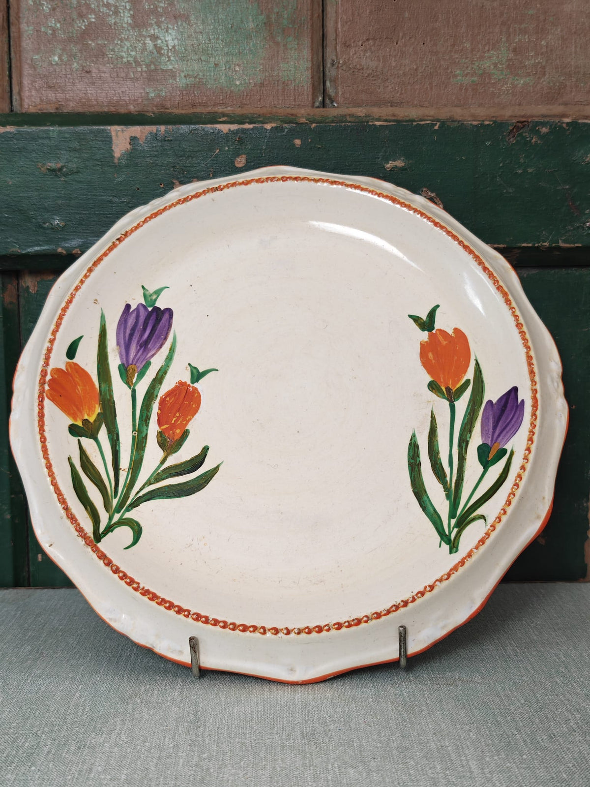 A round off-white ceramic serving platter with scalloped edges, decorated with two painted floral clusters in purple and orange with green stems, and a thin orange beaded border. 