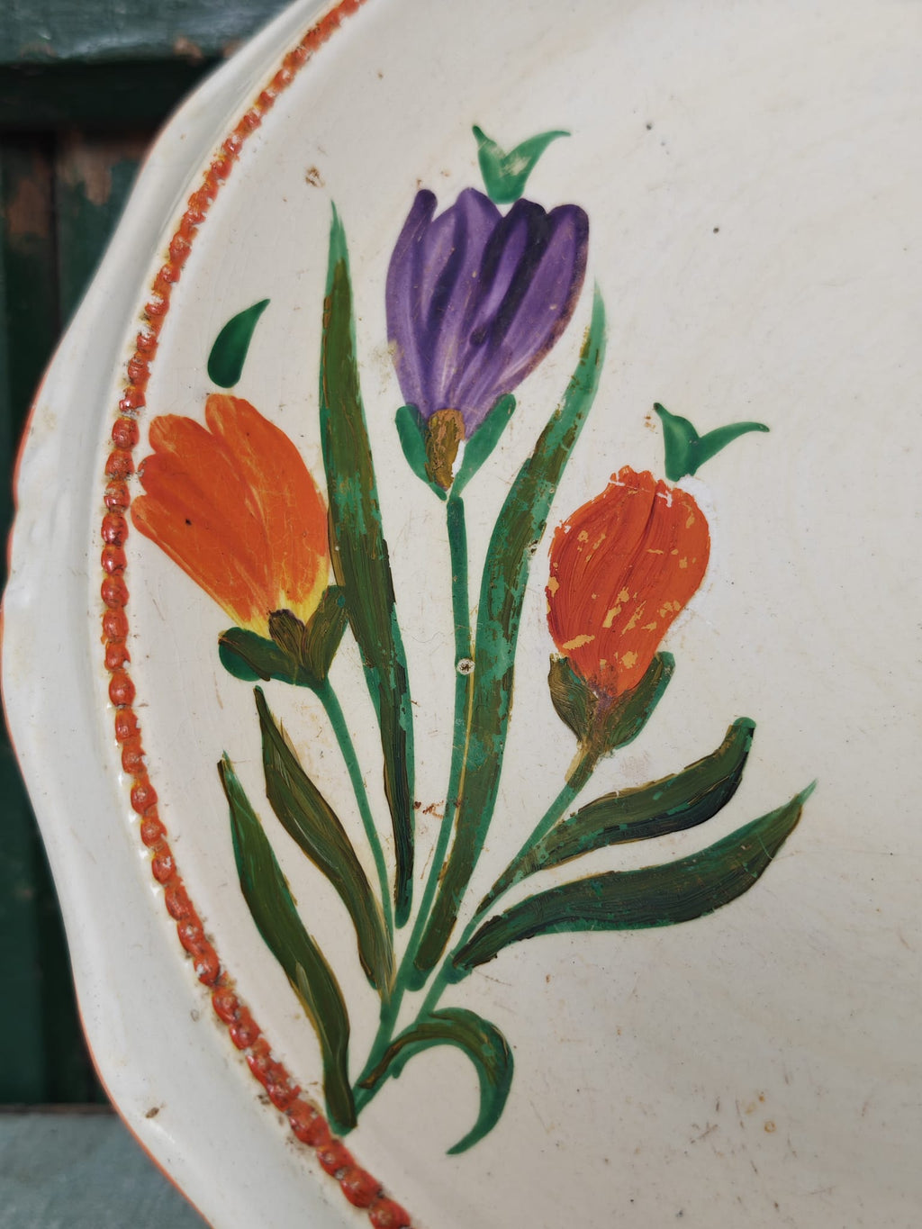 A close-up of a white ceramic plate featuring a hand-painted floral design with orange tulips, a purple flower, green leaves, and a decorative orange beaded border.