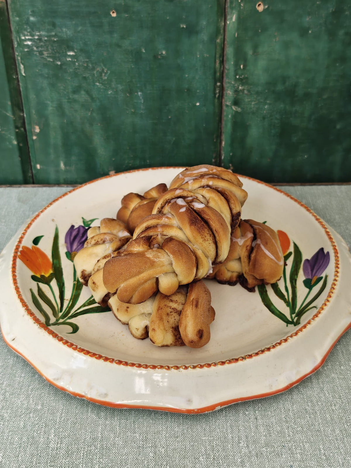 A pile of golden-brown twisted cinnamon rolls or braided bread rolls topped with a glossy glaze, served on a white ceramic plate with a floral design.