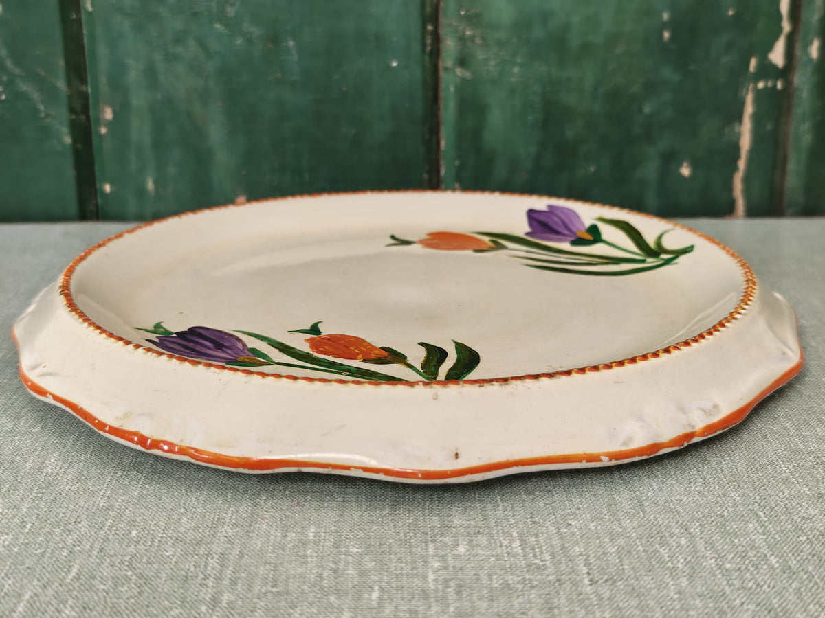 An round ceramic serving platter with a scalloped rim, featuring a hand-painted floral pattern of purple and orange flowers with green leaves. 