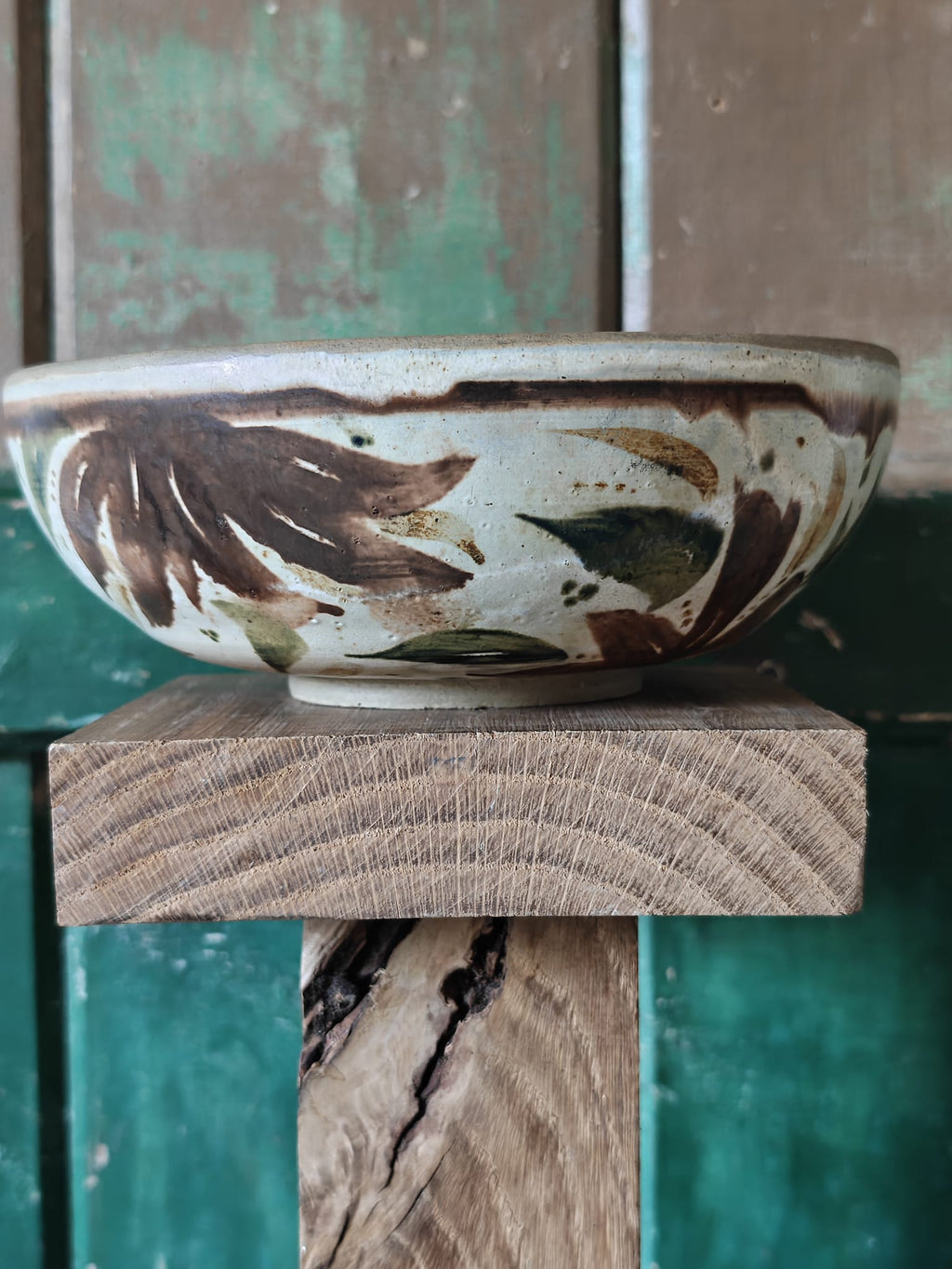 A handmade ceramic bowl with a rustic hand-painted pattern of brown and green leaves on a cream background.