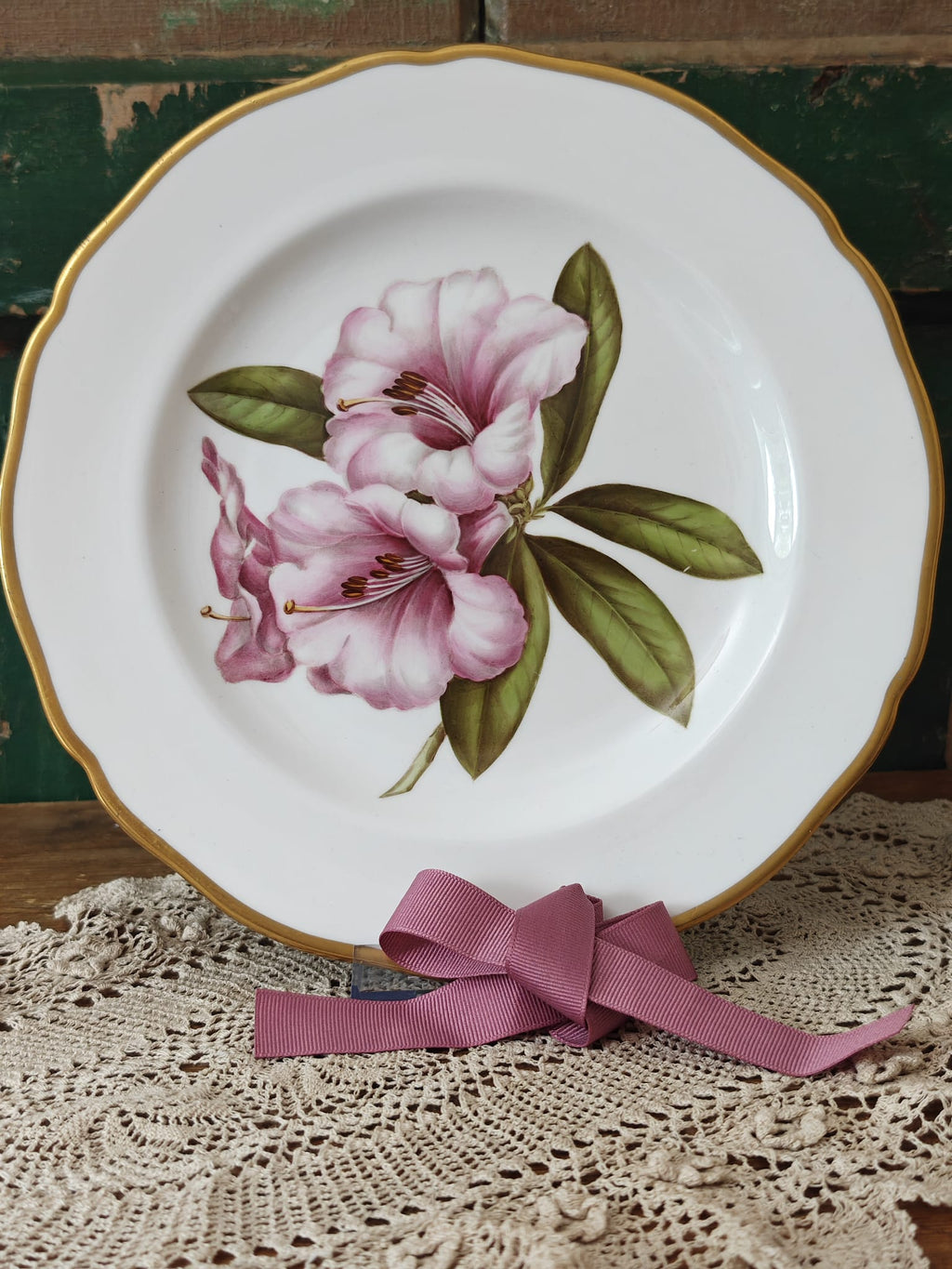 A white ceramic plate with a gold scalloped rim, decorated with pink rhododendron flowers and green leaves. 