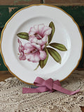 A white ceramic plate with a gold scalloped rim, decorated with pink rhododendron flowers and green leaves. 
