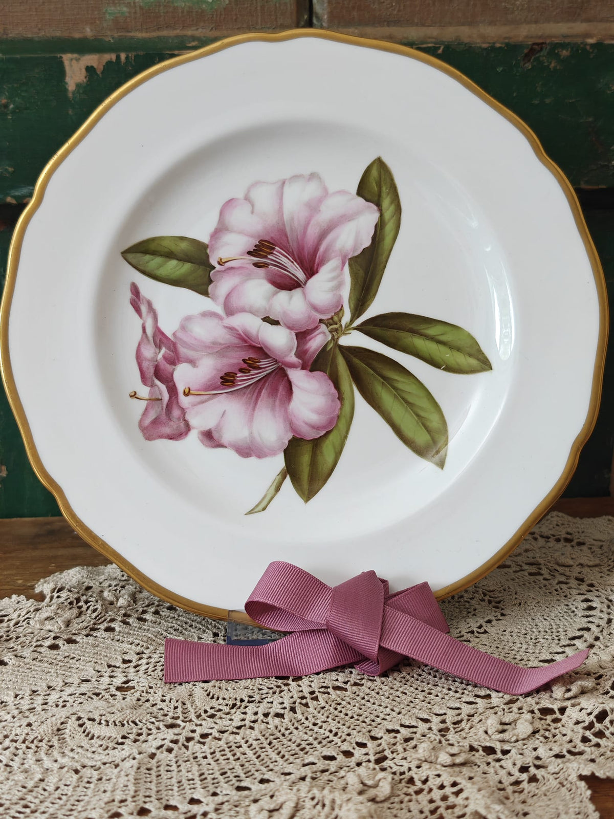 A white ceramic plate with a gold scalloped rim, decorated with pink rhododendron flowers and green leaves. 