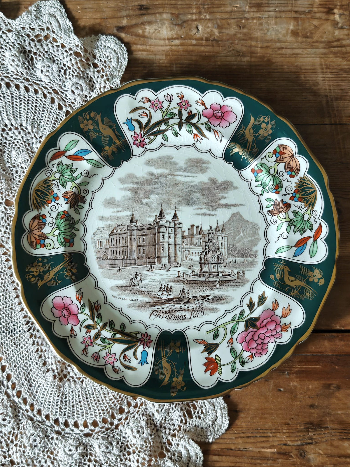 An ornate vintage Christmas plate with a dark green border, pink floral motifs, and a central sepia illustration of a palace.