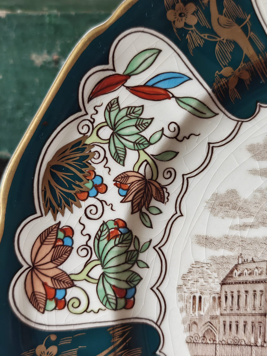 Close-up of an ornate ceramic plate featuring a white ground decorated with teal blue border, gold leaf trim, and detailed botanical motifs.