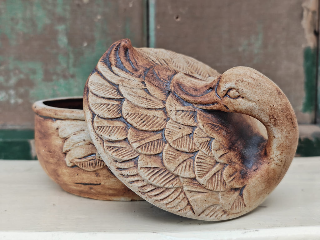 A rustic ceramic duck shaped dish with a matching lid, featuring detailed feather carvings and a natural earthy brown and beige glaze.