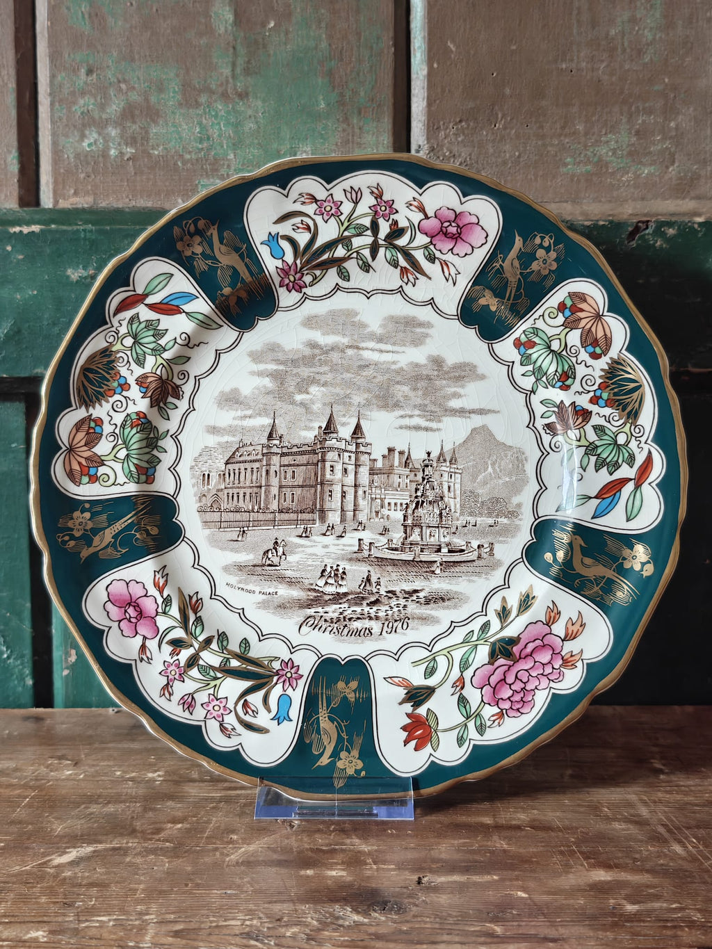 An ornate round Christmas plate with a detailed illustration of a castle and boats in the center, surrounded by a decorative border with pink flowers, birds, and foliage. 