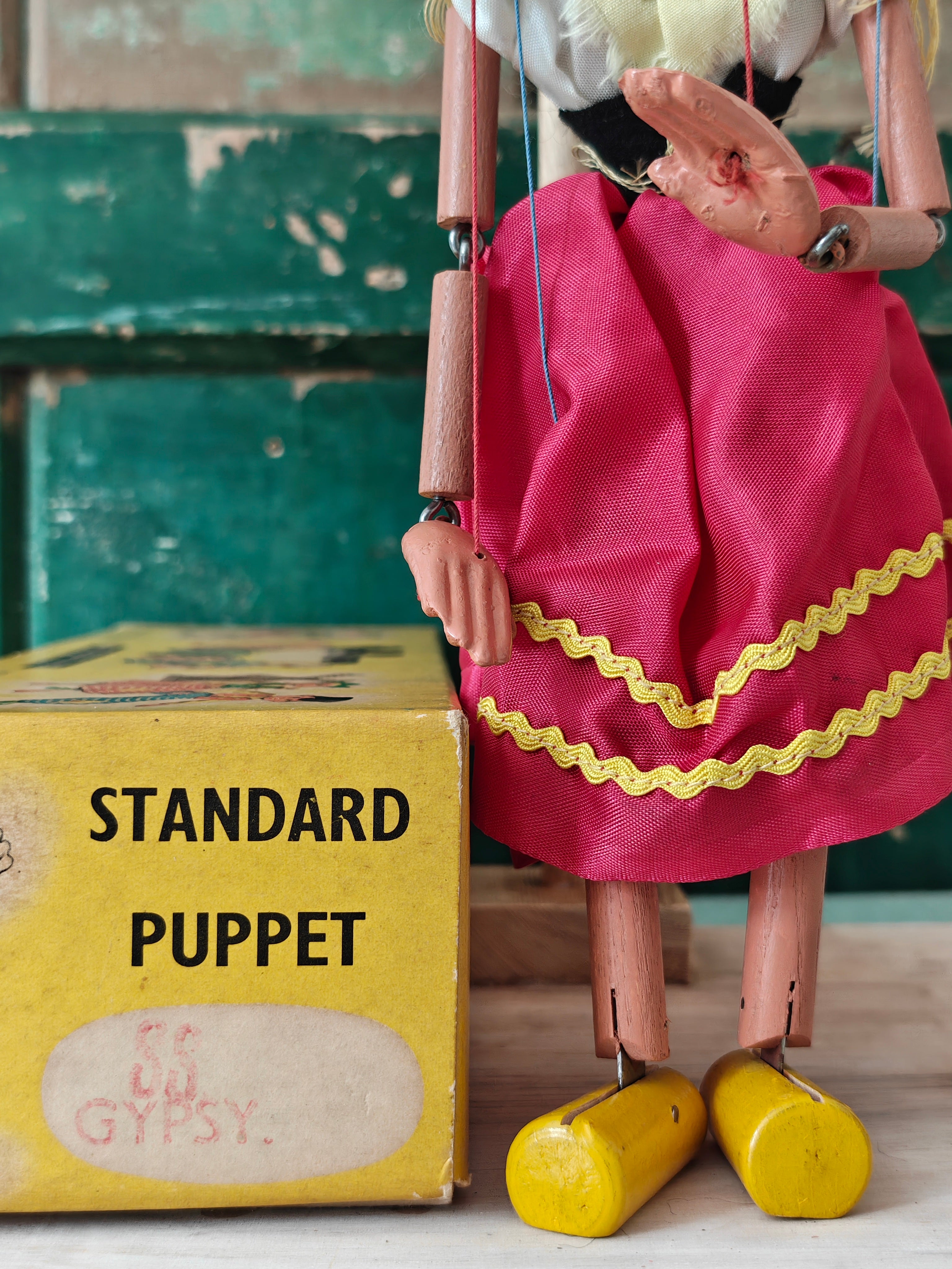 Wooden puppet with a pink dress and yellow shoes next to a box labeled 'Standard Puppet' on a wooden floor.