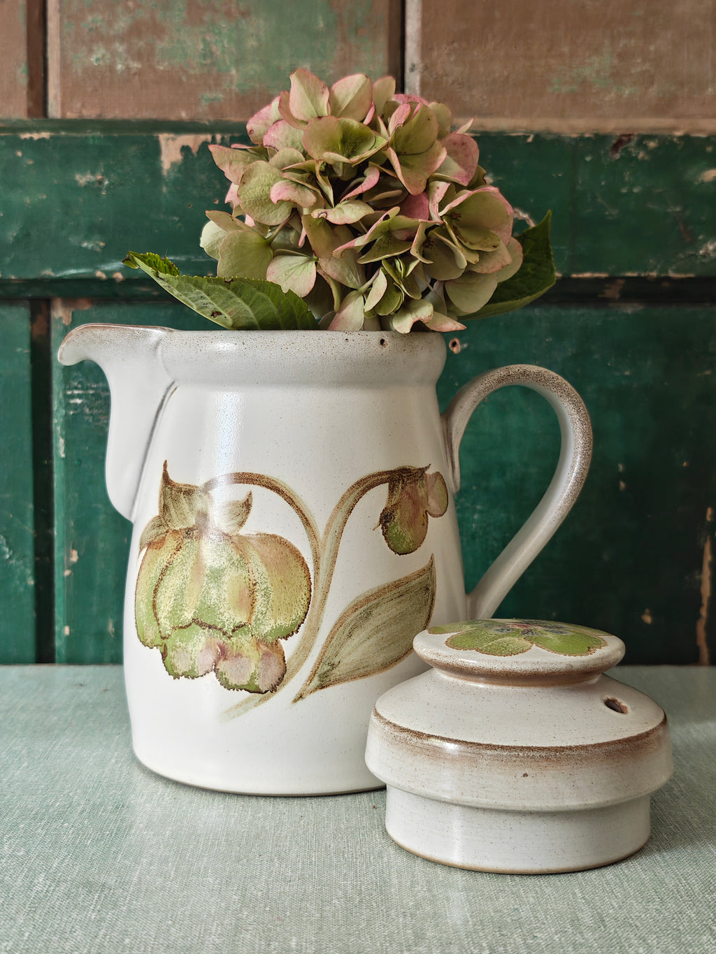 A vintage ceramic coffee pot with a cream-colored glaze, featuring a painted green and pink magnolia flower design. The pot has a matching lid, a curved handle, and a spout.