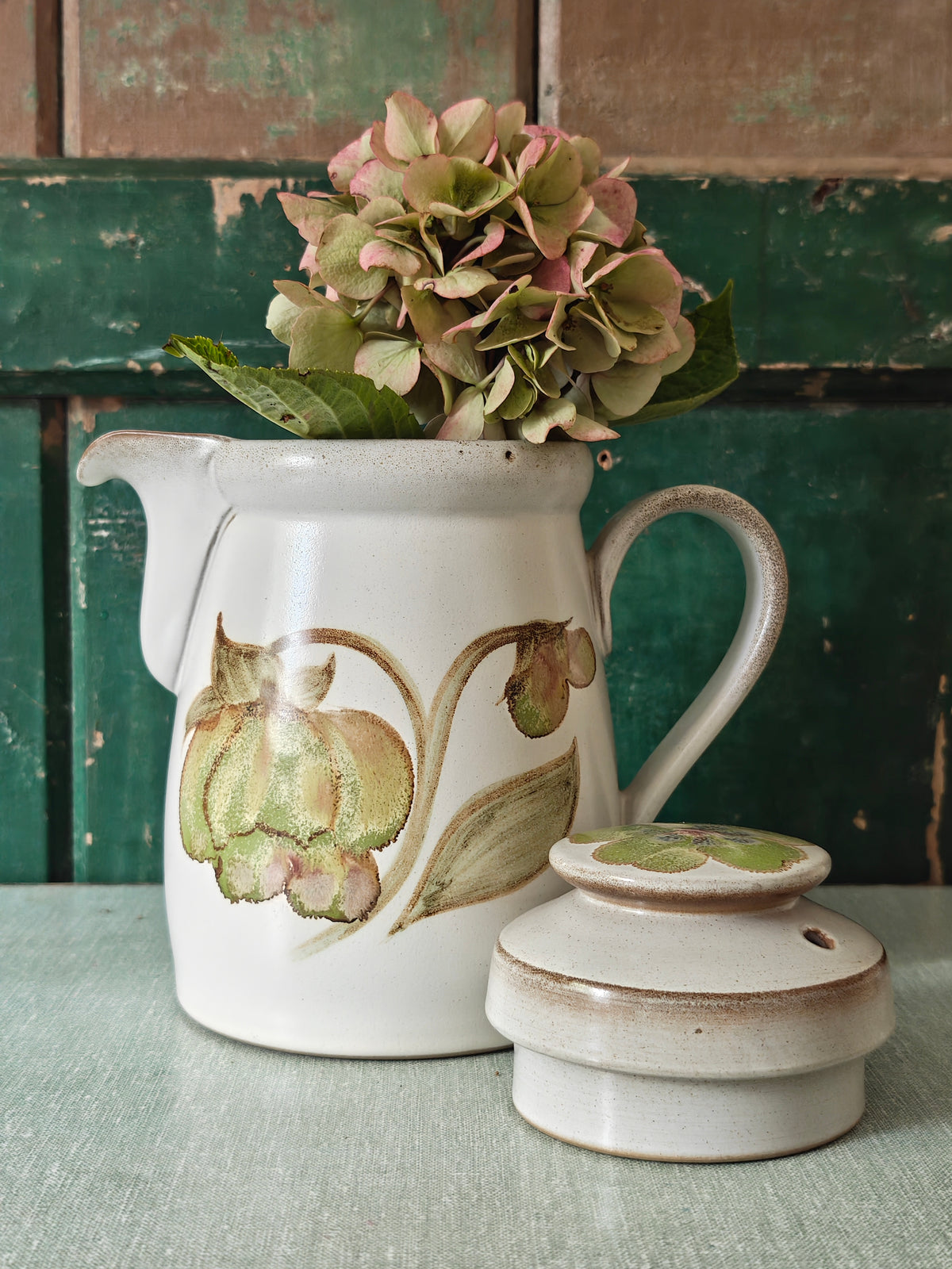 A vintage ceramic coffee pot with a cream-colored glaze, featuring a painted green and pink magnolia flower design. The pot has a matching lid, a curved handle, and a spout.