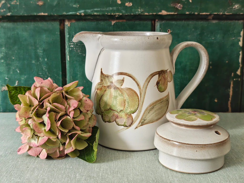 A vintage ceramic coffee pot with a cream-colored glaze, featuring a painted green and pink magnolia flower design. The pot has a matching lid, a curved handle, and a spout.