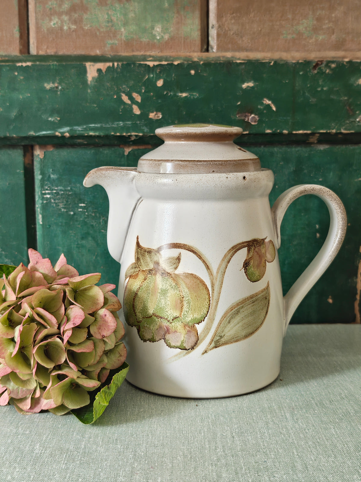 A vintage ceramic coffee pot with a cream-colored glaze, featuring a painted green and pink magnolia flower design. The pot has a matching lid, a curved handle, and a spout.
