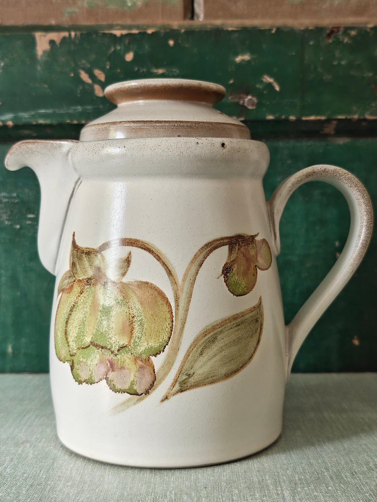 A vintage ceramic coffee pot with a cream-colored glaze, featuring a painted green and pink magnolia flower design. The pot has a matching lid, a curved handle, and a spout.
