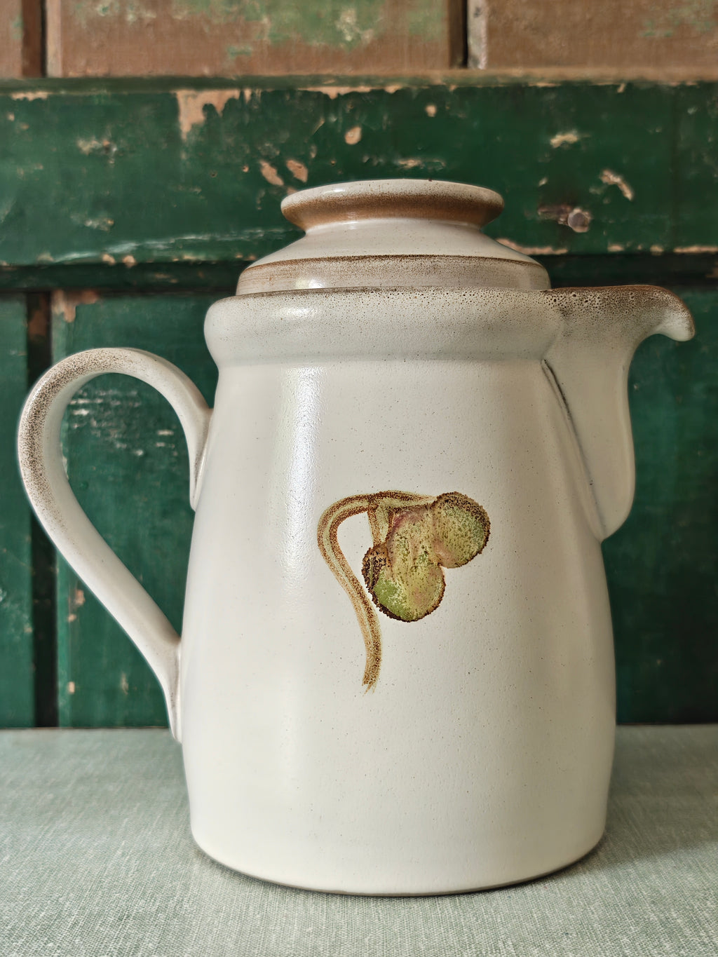 A vintage ceramic coffee pot with a cream-colored glaze, featuring a painted green and pink magnolia flower design. The pot has a matching lid, a curved handle, and a spout.
