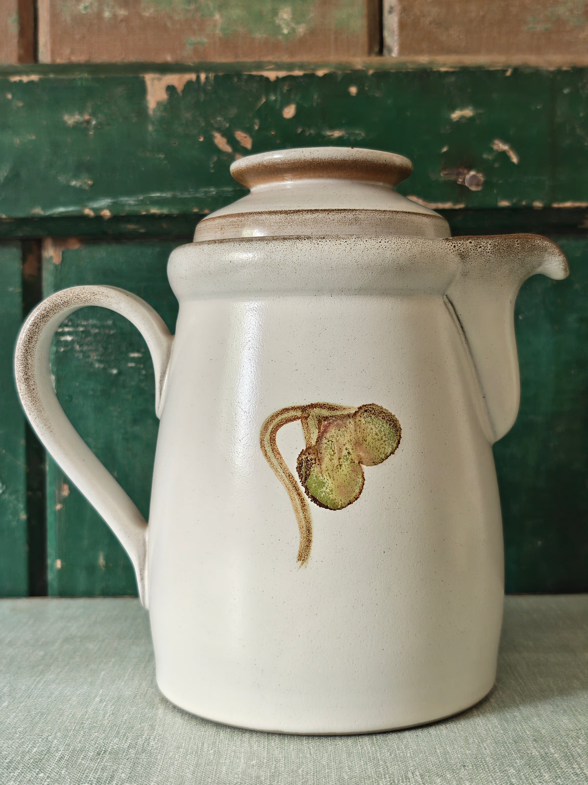 A vintage ceramic coffee pot with a cream-colored glaze, featuring a painted green and pink magnolia flower design. The pot has a matching lid, a curved handle, and a spout.