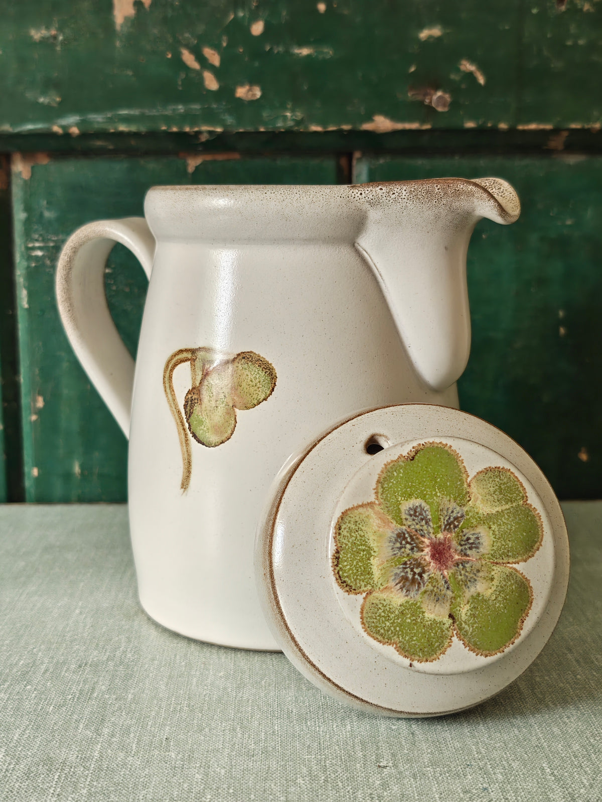 A vintage ceramic coffee pot with a cream-colored glaze, featuring a painted green and pink magnolia flower design. The pot has a matching lid, a curved handle, and a spout. The photo shows the lid leaning against the side of the coffee pot.