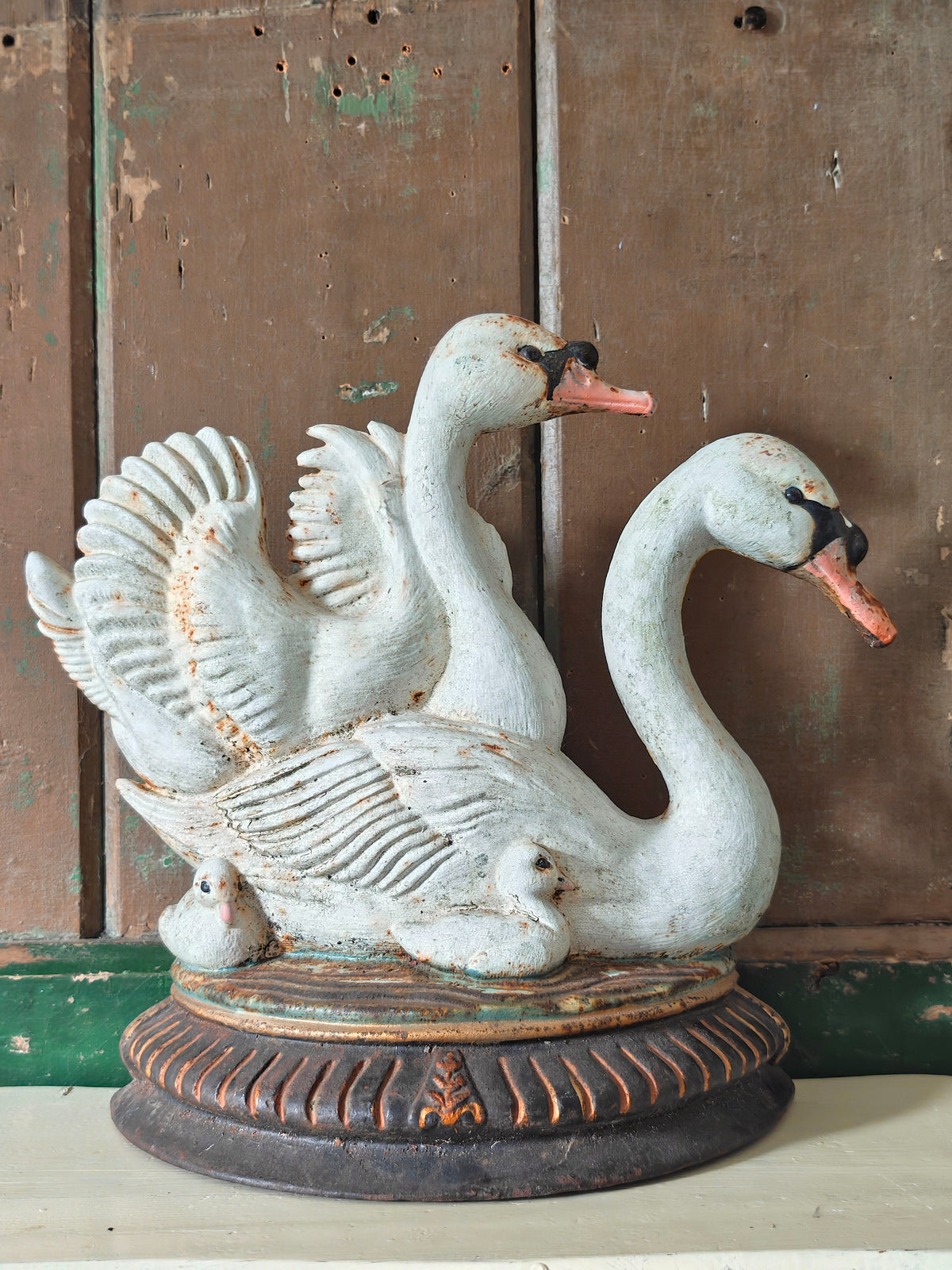 A decorative cast iron doorstop modelled as two swans and cygnets on a round pedestal base. The swans are painted in a distressed off-white finish with orange beaks and black facial details.