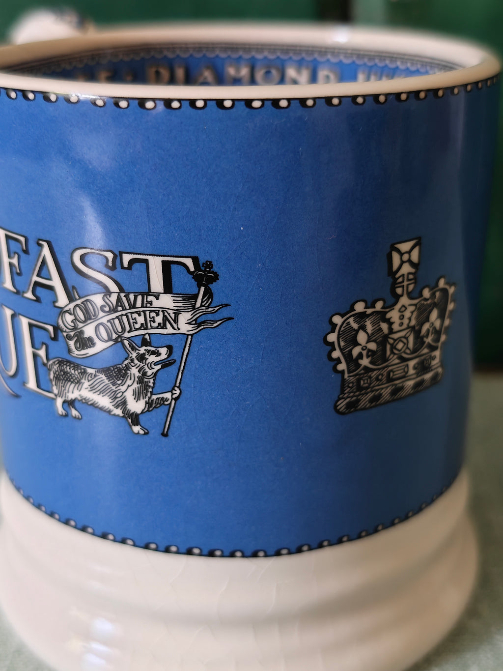 A close-up of a ceramic mug featuring a blue upper body with white and black printed graphics, including a crowned emblem and partial text, and a white lower body.