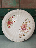A vintage scalloped ceramic plate with a distressed white glaze, decorated with pink floral and green leaf motifs.