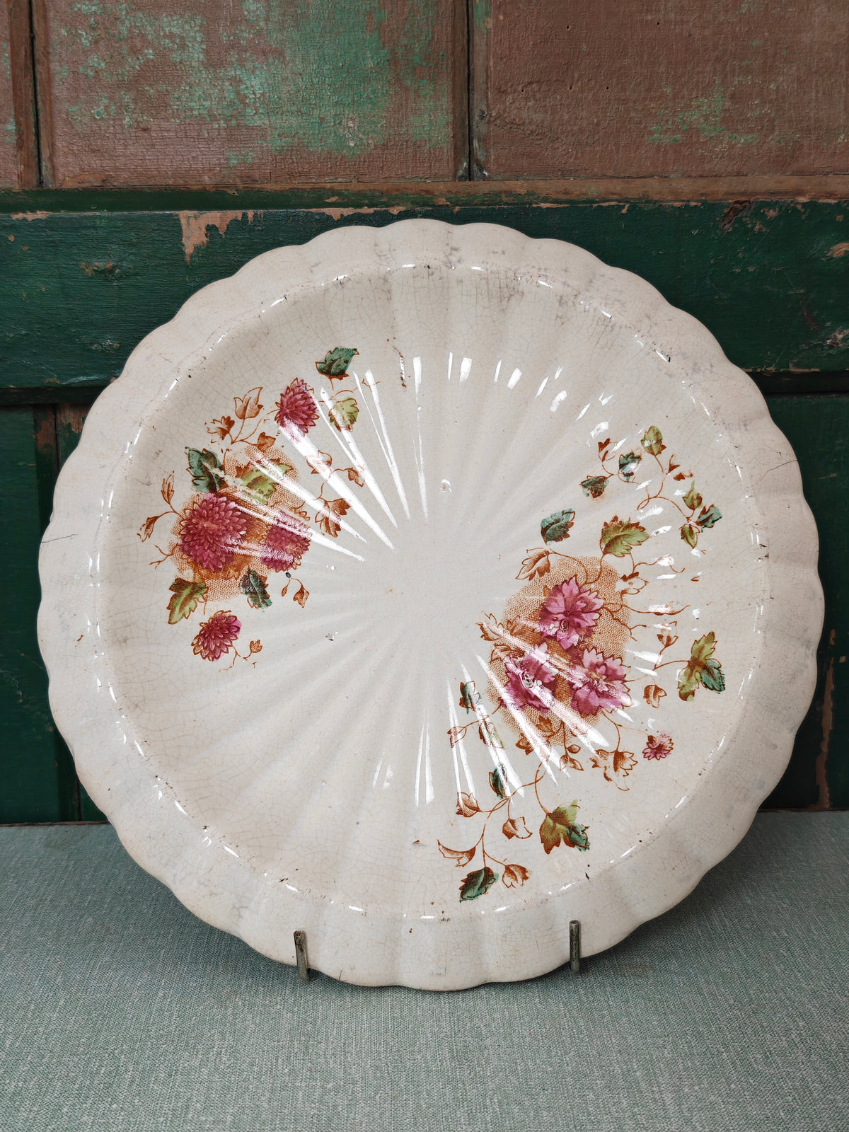 A vintage scalloped ceramic plate with a distressed white glaze, decorated with pink floral and green leaf motifs.