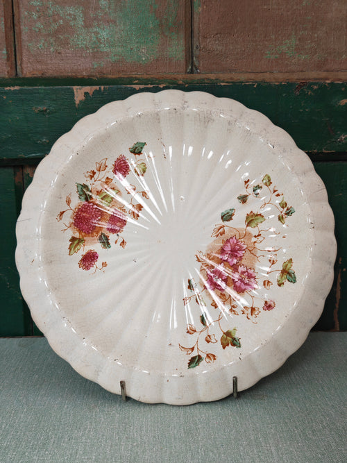 A vintage scalloped ceramic plate with a distressed white glaze, decorated with pink floral and green leaf motifs.