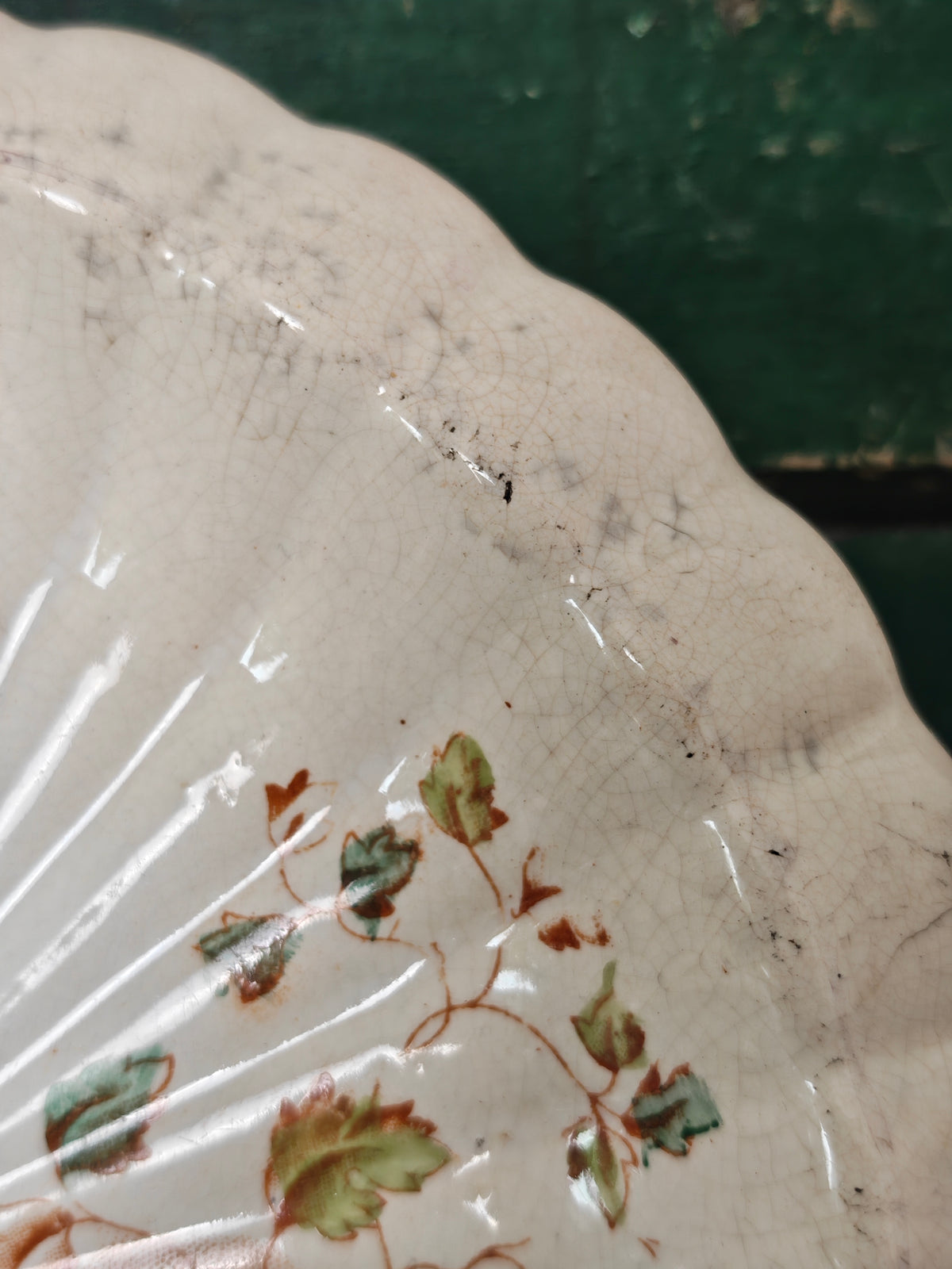 A close-up of a vintage ceramic plate with a scalloped edge, featuring a glossy off-white glaze with a delicate green and brown floral vine pattern. 