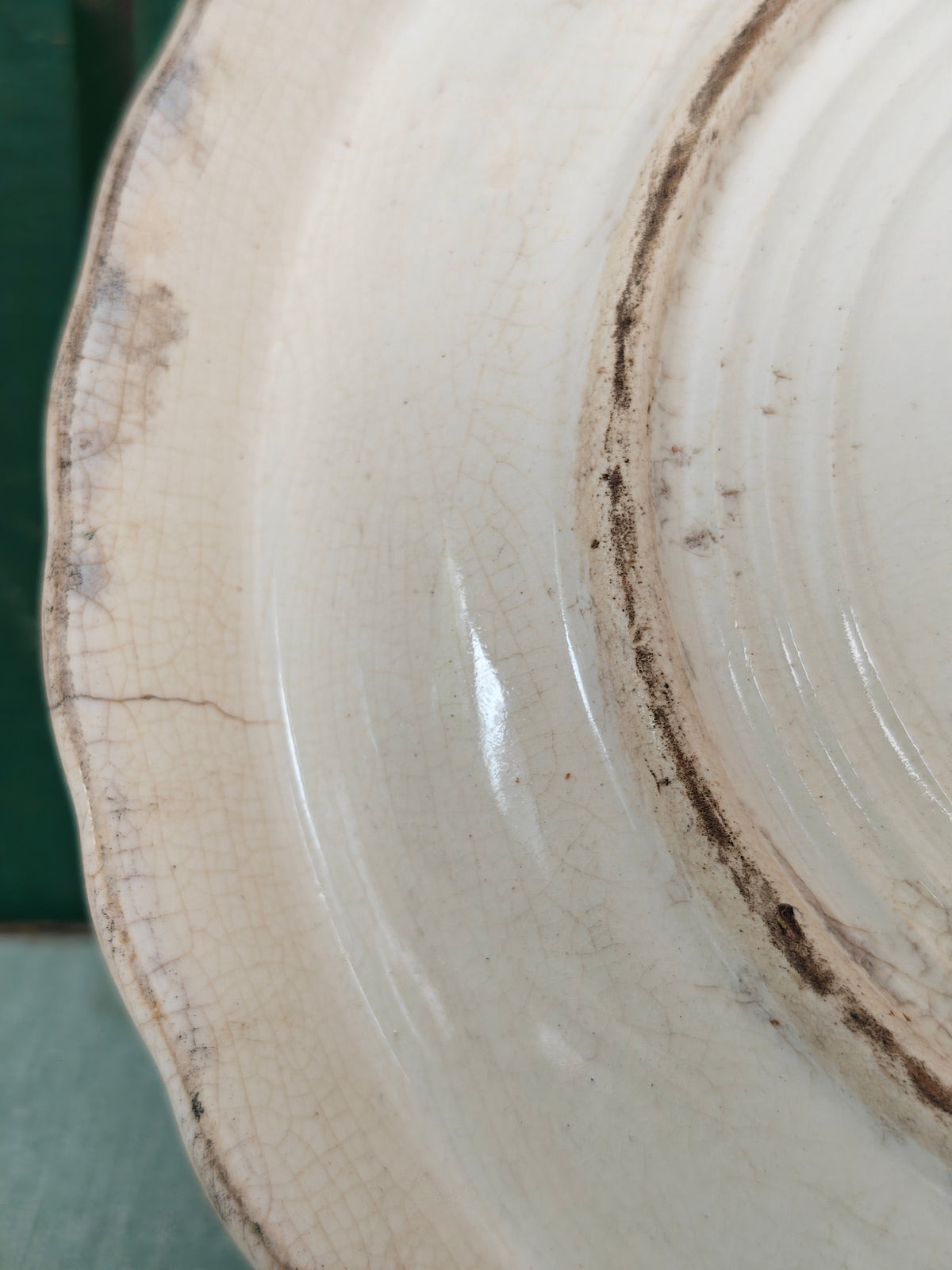 Close-up of the back of a vintage ceramic plate or dish with a scalloped rim, featuring a cream-colored glaze with prominent crackle patterns and dark brown staining along the edge.