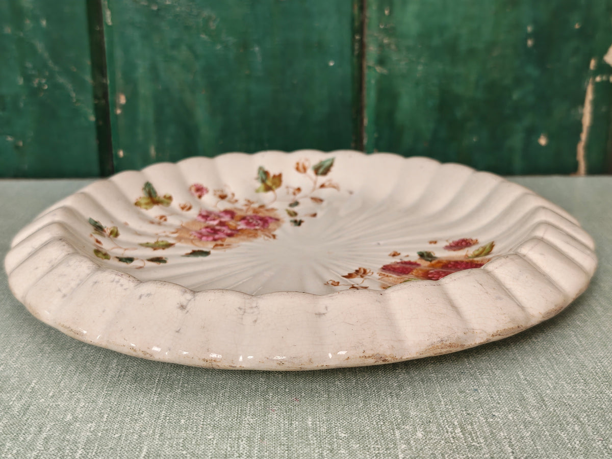 A vintage scalloped ceramic plate with a distressed white glaze, decorated with pink floral and green leaf motifs.