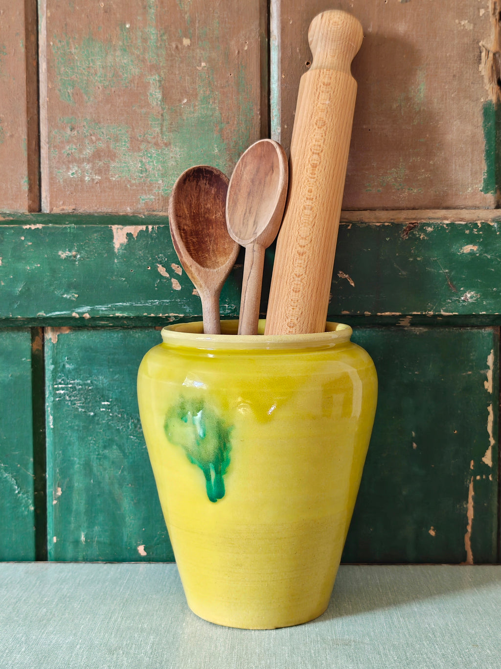A yellow glazed ceramic jar with a green drip glaze design, displayed with kitchen utensils