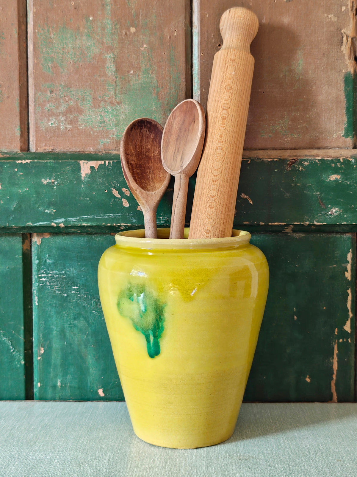 A yellow glazed ceramic jar with a green drip glaze design, displayed with kitchen utensils